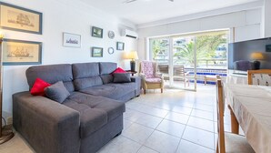 A living room with a grey sofa, a red pillow, a chair with a floral cover, a TV, and a balcony with a view of palm trees and a pool. - Apartment Navio by Interhome (Aguadulce)