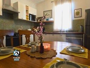 A dining table with a vase of flowers, salt and pepper shakers, and a candle. There is a chair and a dining table set with plates and bowls. The kitchen in the background has cabinets and a microwave.
