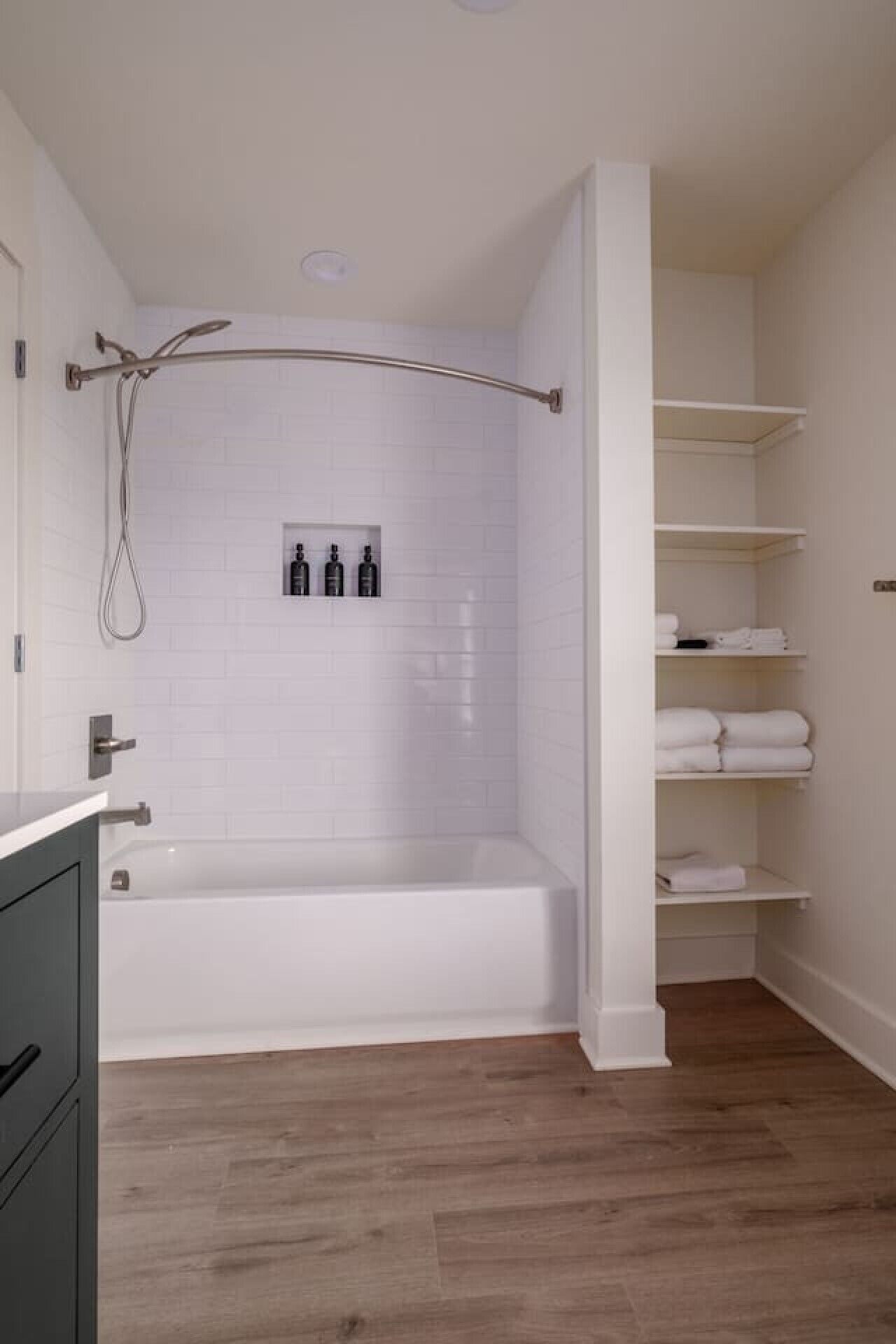 Une salle de bain moderne avec une baignoire, des murs recouverts de carreaux blancs, un plancher en bois et une étagère intégrée où sont rangées les serviettes.