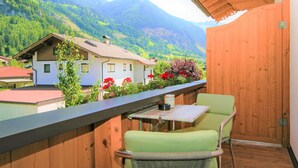 A balcony with a wooden railing, a table, and two chairs. A house with a red roof and a garden with red flowers is visible in the background. - Paradiso by Interhome (Ramsau im Zillertal)