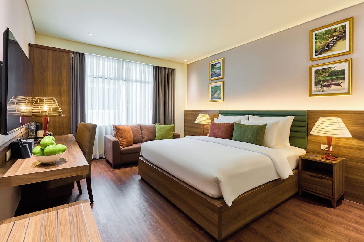 Ein Hotelzimmer mit einem großen Bett, einem Schreibtisch mit Obstschale, einem Fernseher und einem Fenster mit Vorhängen.