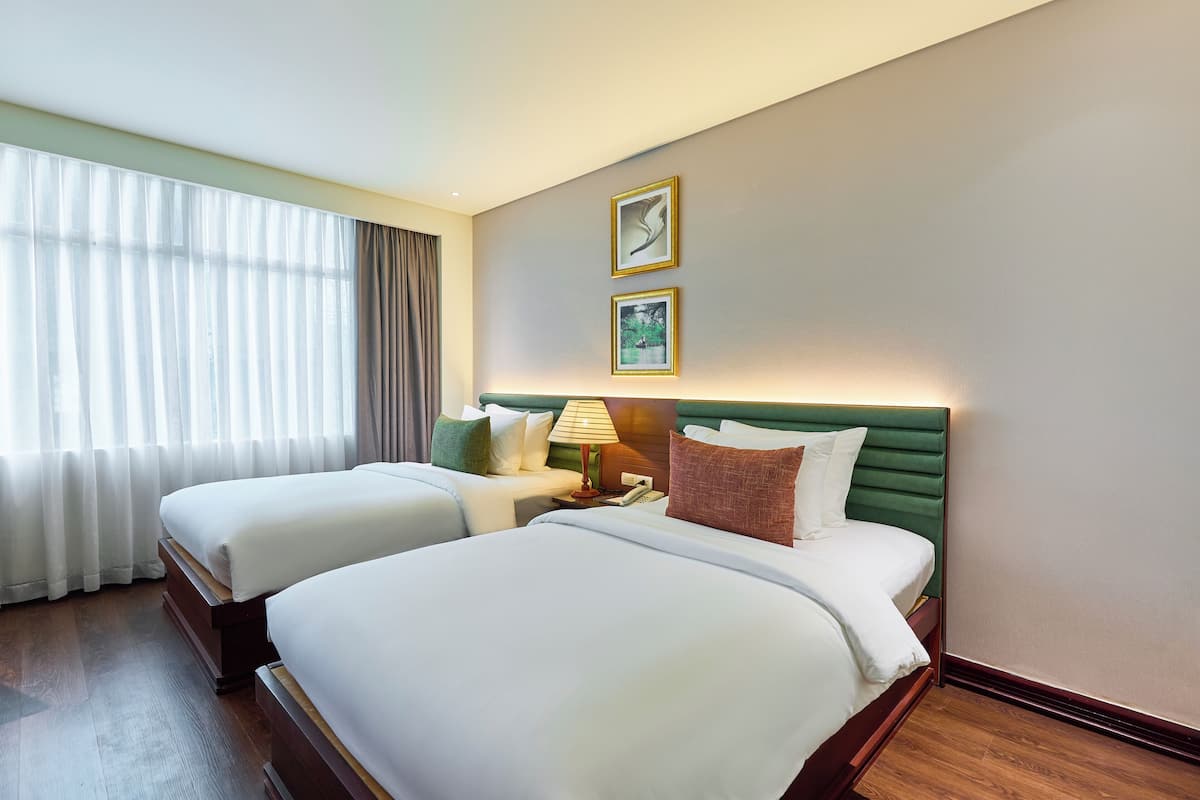 Ein Hotelzimmer mit zwei Betten, einem Nachttisch, einer Lampe und einem großen Fenster mit Vorhängen.