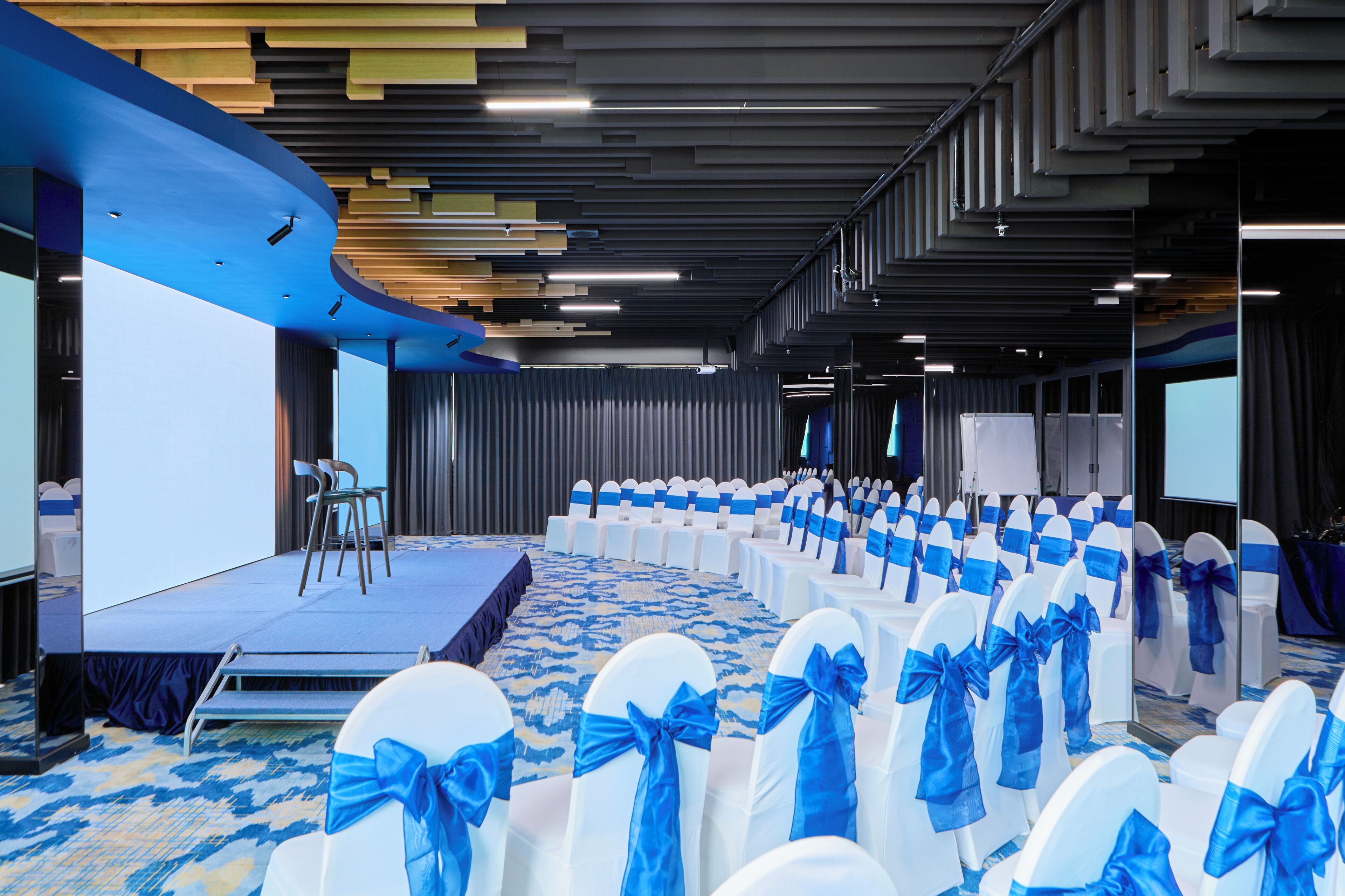 A conference room with rows of white chairs with blue sashes, a stage, and a carpeted floor.