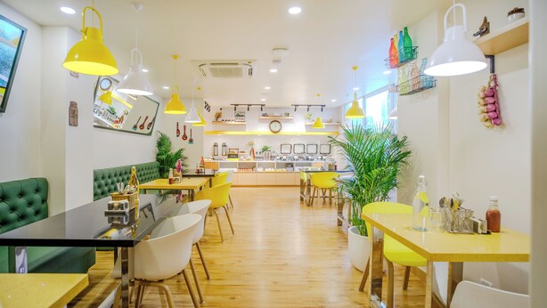 A restaurant interior with wooden flooring, yellow and white tables, green and white chairs, and yellow pendant lights. - Bloom Hotel - HAL Old Airport Rd (Bengaluru)