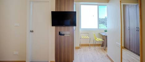 Un dormitorio con cama, televisor montado en un panel de madera, escritorio con silla y ventana con vista a follaje.