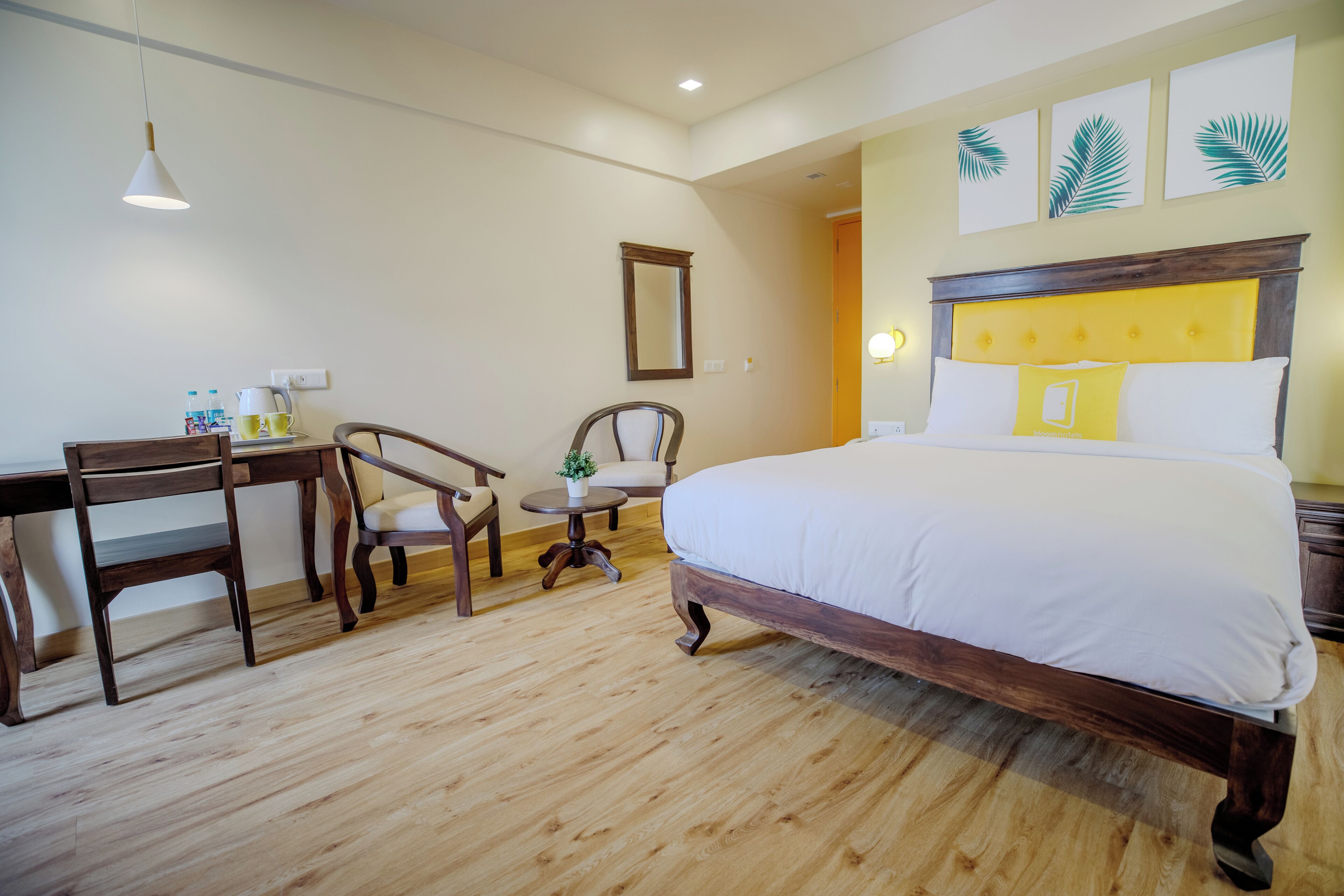 Habitación de hotel con una cama de madera, un escritorio, dos sillas, una mesita con una planta y un espejo en la pared.