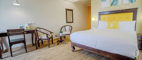 Habitación de hotel con una cama de madera, un escritorio, dos sillas, una mesita con una planta y un espejo en la pared.