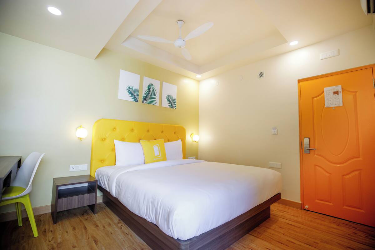 Une chambre d’hôtel avec un lit, un bureau, une chaise, un ventilateur de plafond et une porte orange.