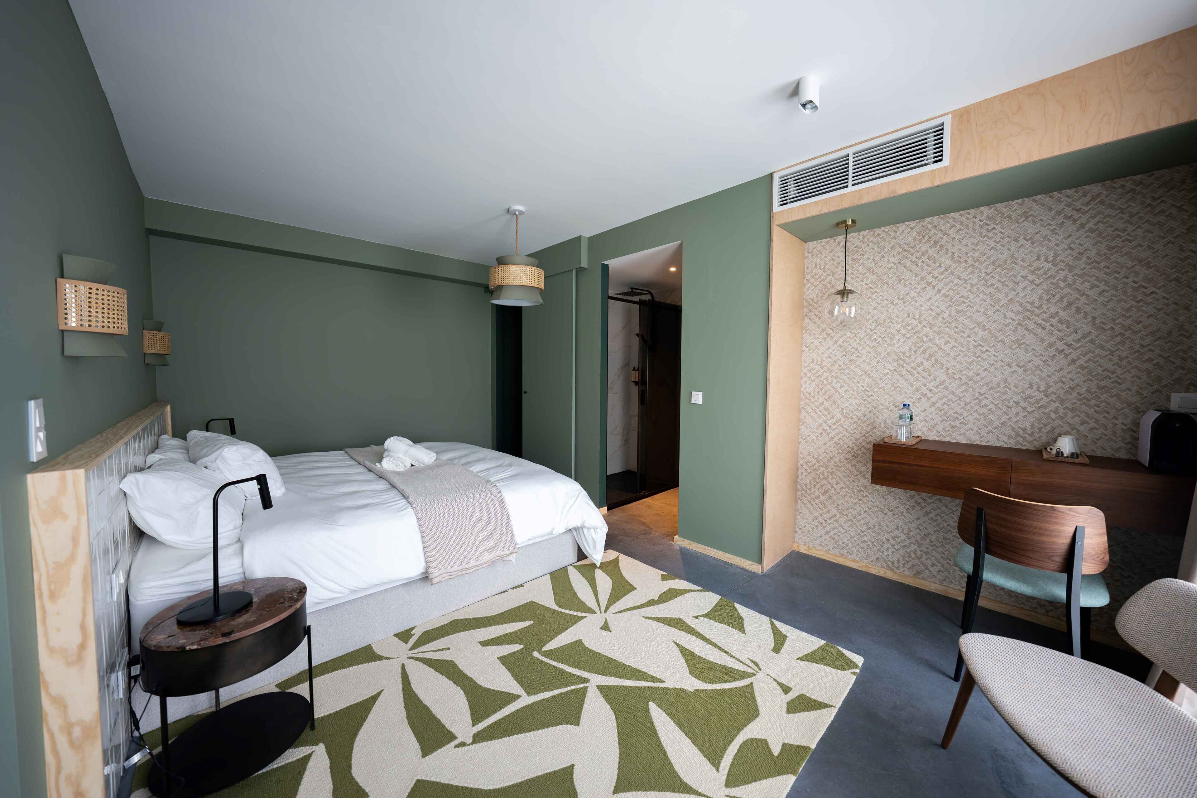 A modern hotel room with a bed, bedside table, chair, and a desk area.
