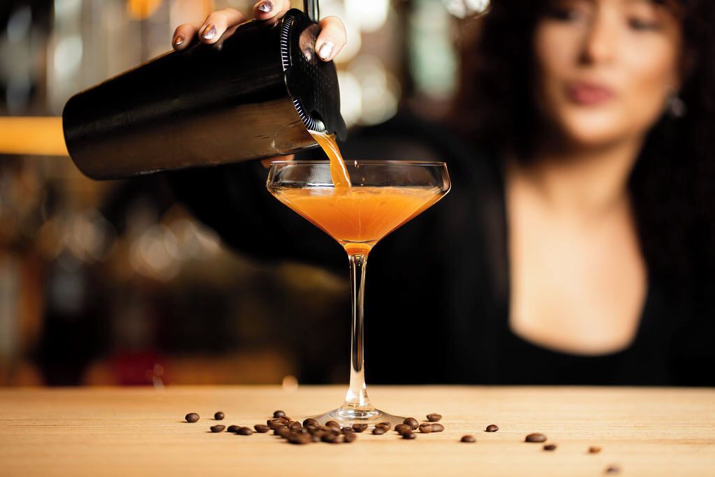 A cocktail being poured into a martini glass on a wooden surface with scattered coffee beans.