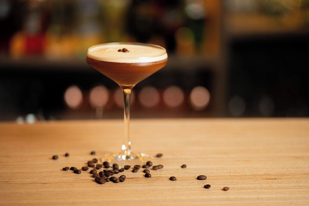 Una copa de martini con una espuma en la parte superior, sobre una superficie de madera, rodeada de granos de café esparcidos.