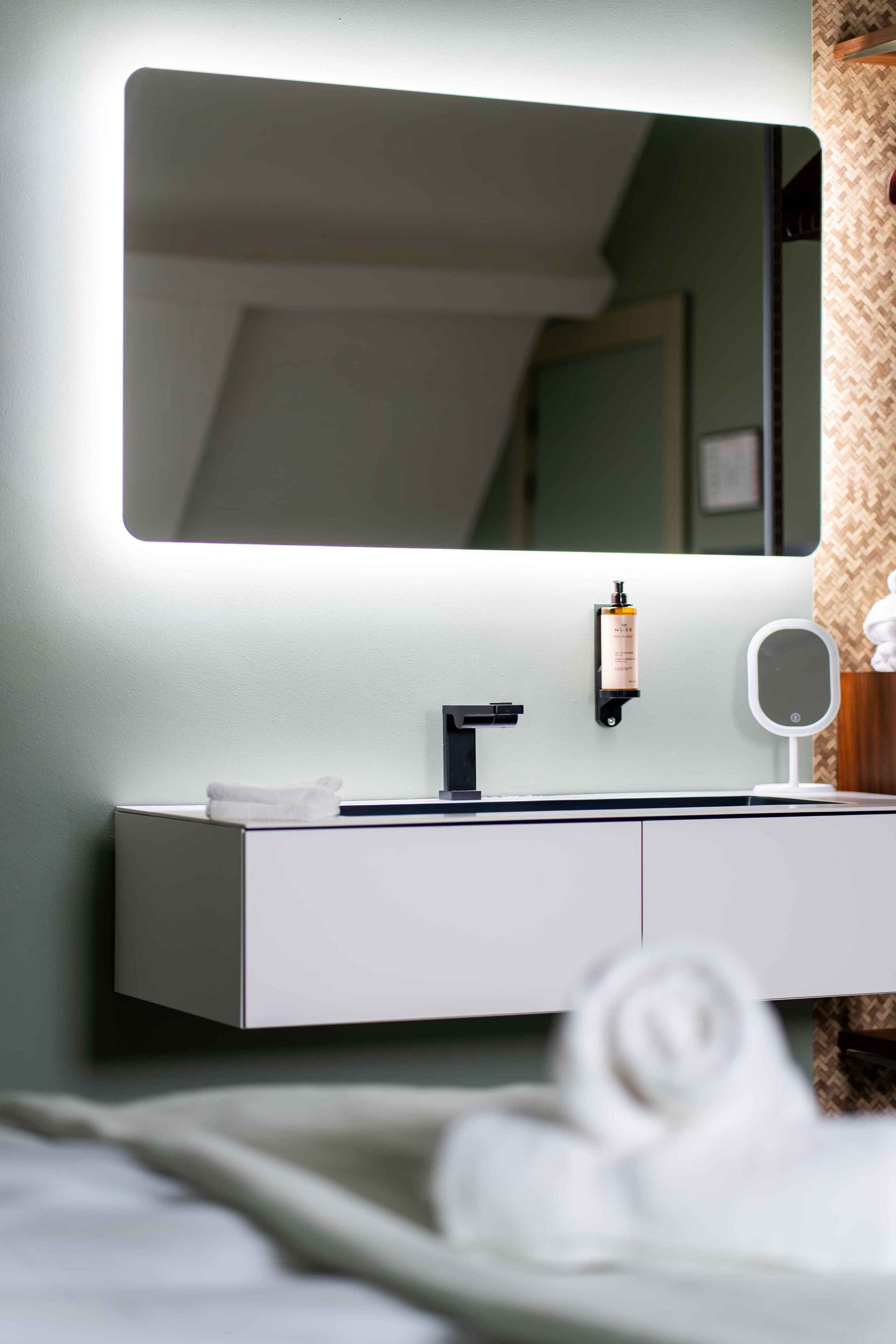 A modern bathroom with a large mirror, a towel on the counter, and a bottle of shampoo.