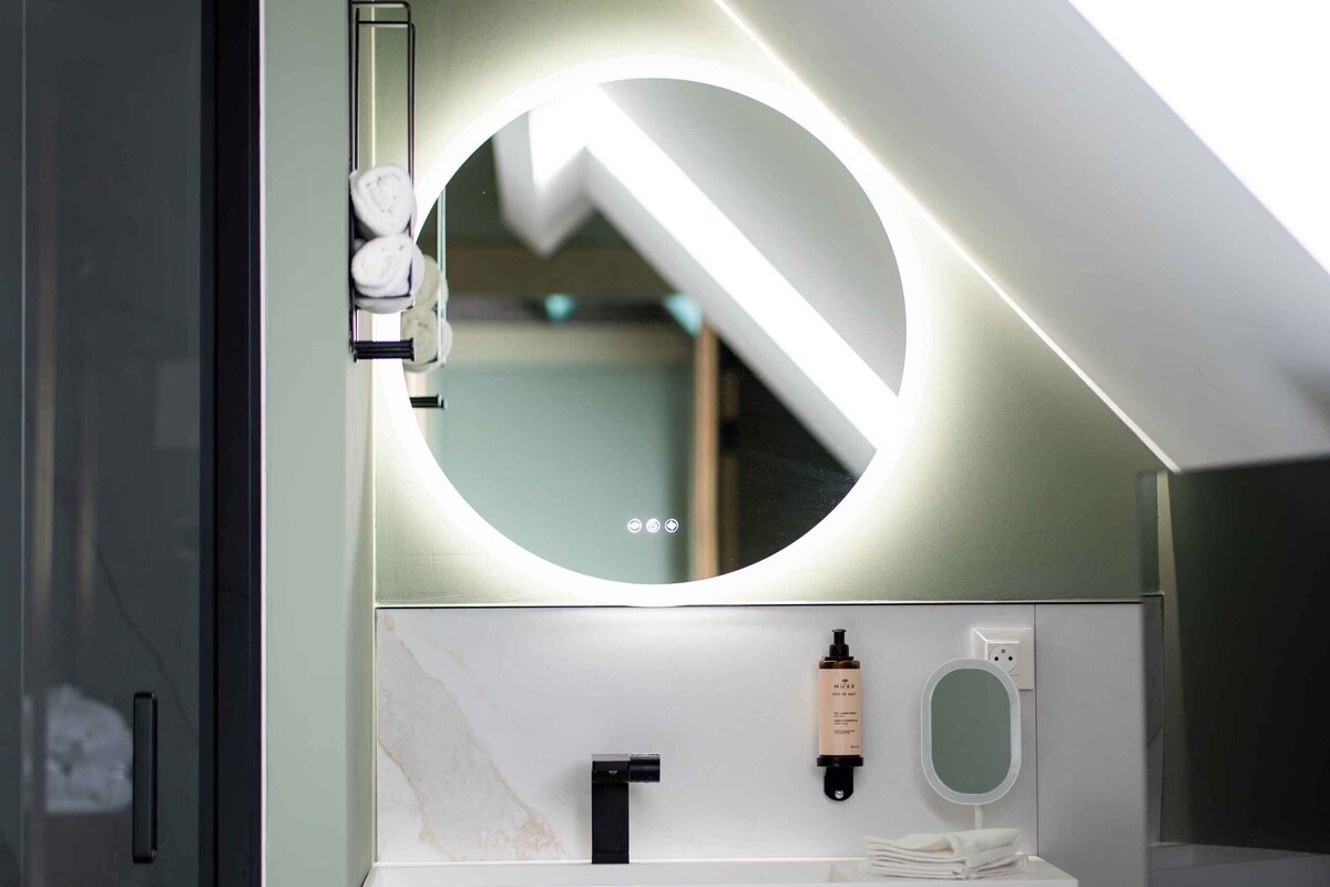 A modern bathroom with a white sink, a round mirror, and a towel dispenser.