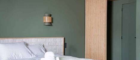 Una cama bien hecha con ropa blanca, cabecera con diseño geométrico y una lámpara fijada a la pared.