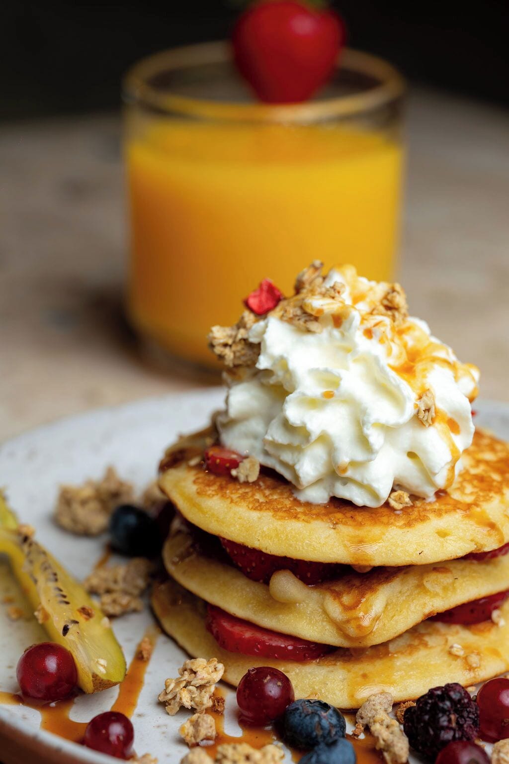 Una pila de panqueques cubiertos de crema batida y frutos rojos, acompañada de un vaso de jugo de naranja.