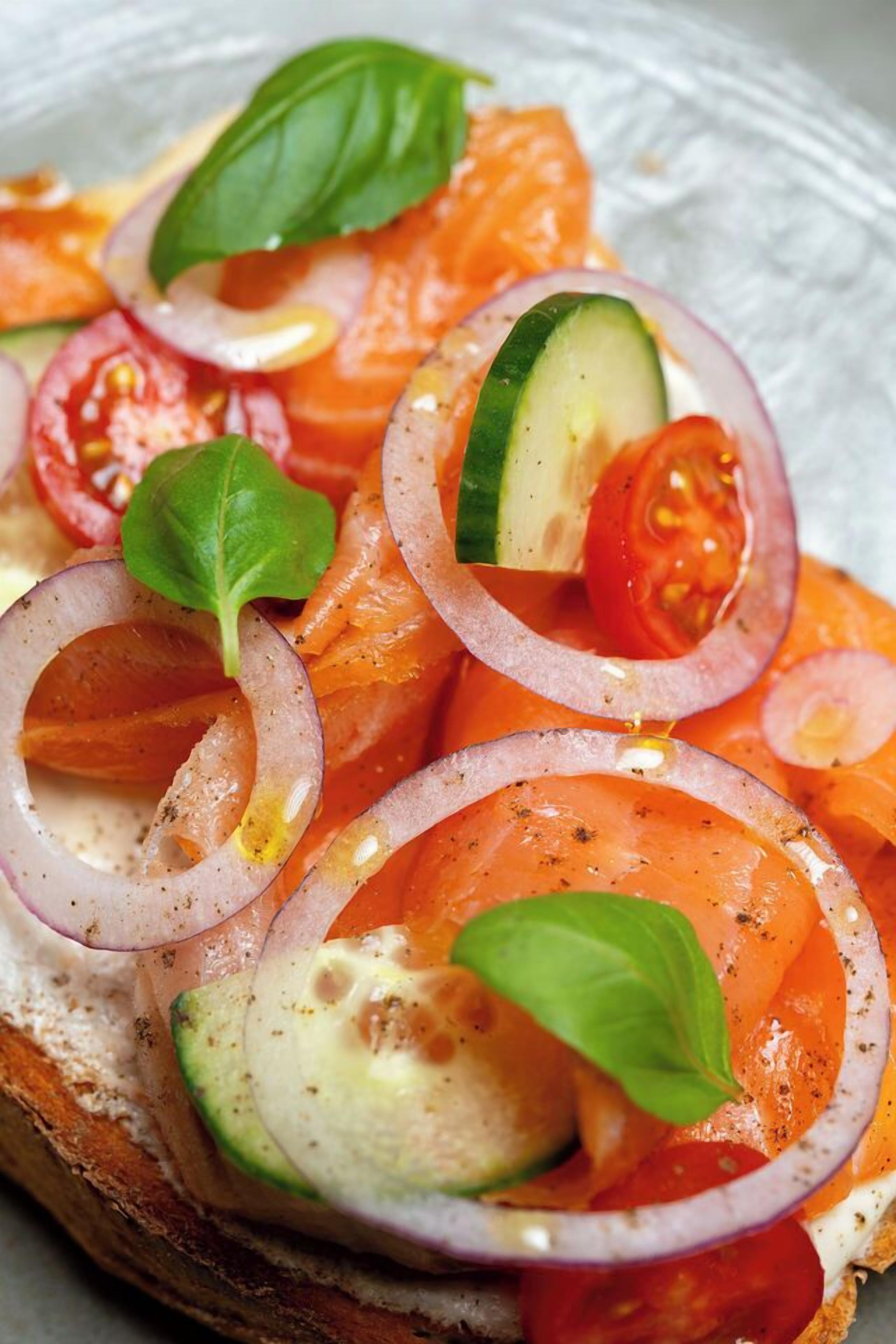 A close-up of a sandwich with smoked salmon, cucumber, tomato, and onion rings, garnished with basil leaves.