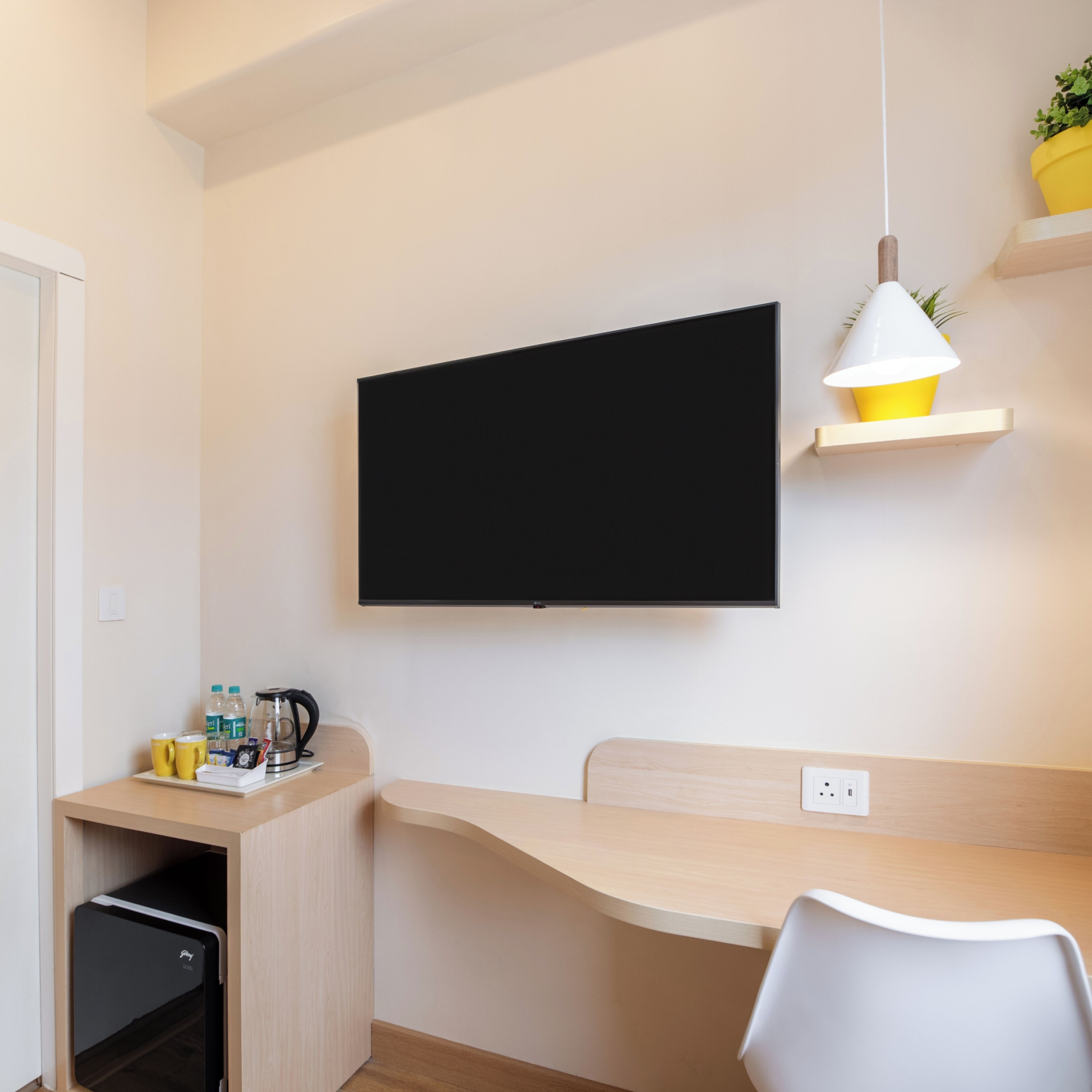 A modern room with a flat-screen TV mounted on the wall, a wooden desk, a white chair, a small shelf with a yellow lamp, and a coffee maker on the desk.