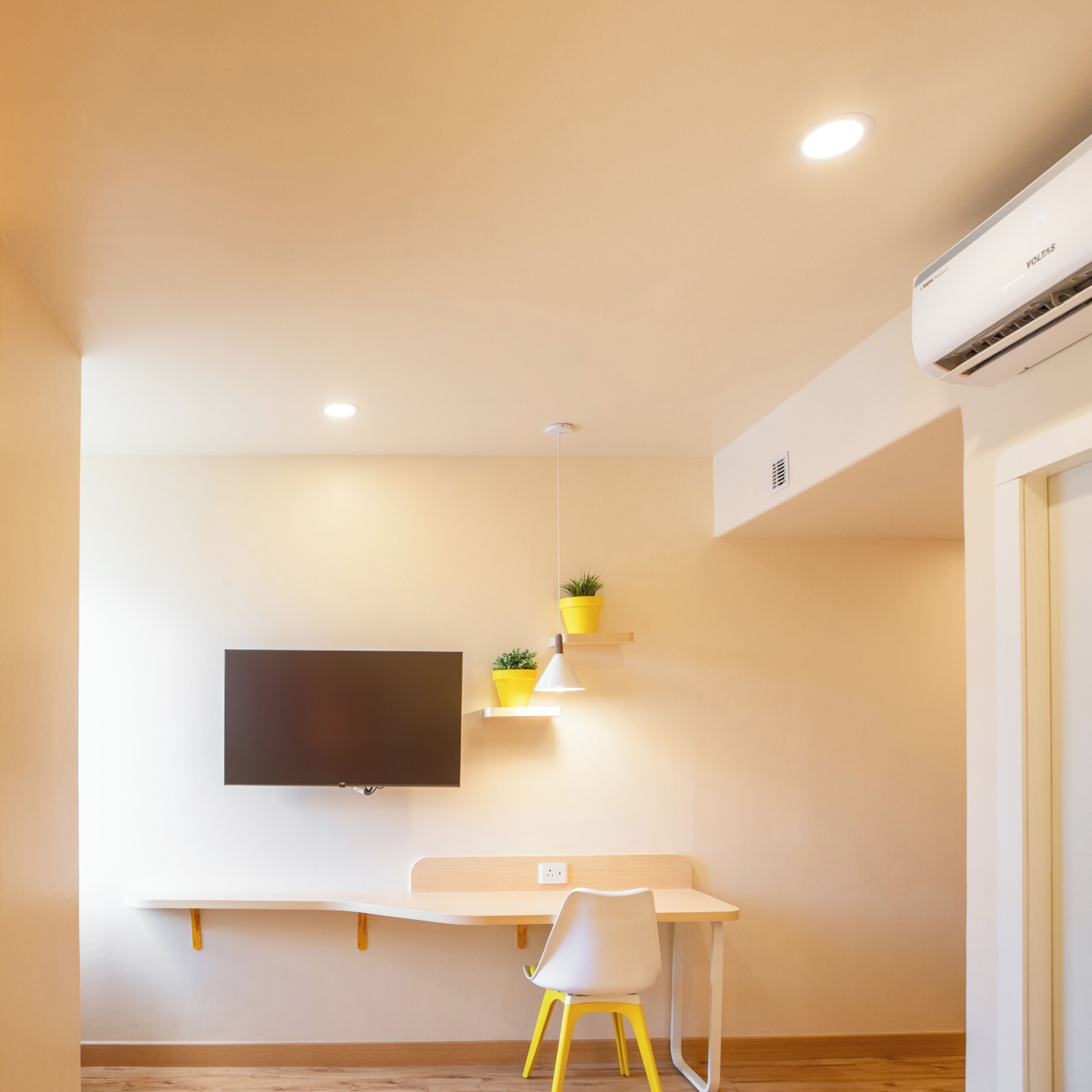 A modern room with a flat-screen TV mounted on the wall, a desk with a chair, and a small potted plant on a shelf.