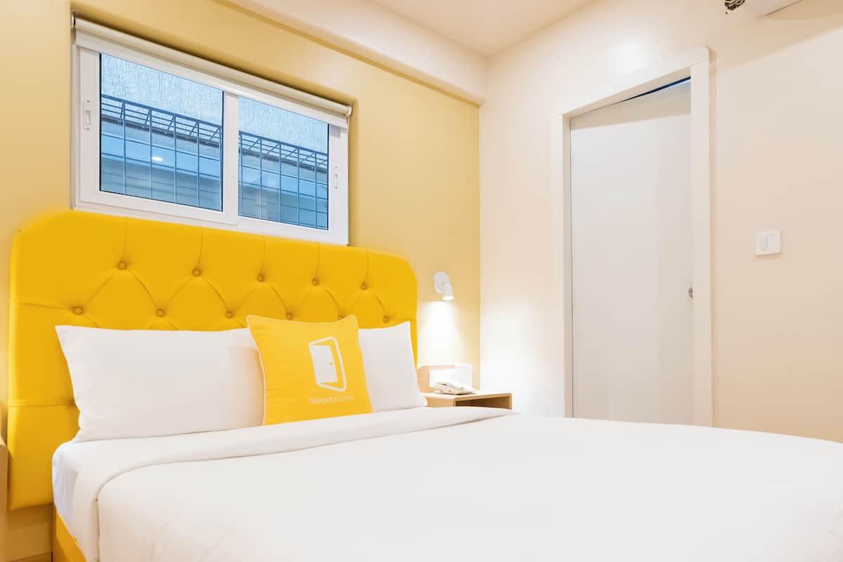 Quarto de hotel com cabeceira amarela, roupa de cama branca e mesa de cabeceira com abajur.