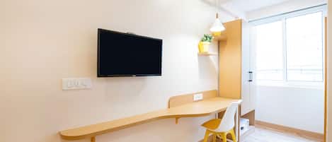 Une chambre moderne dotée d’un téléviseur à écran plat fixé au mur, d’un bureau en bois, d’une chaise jaune et d’une fenêtre laissant entrer la lumière naturelle.