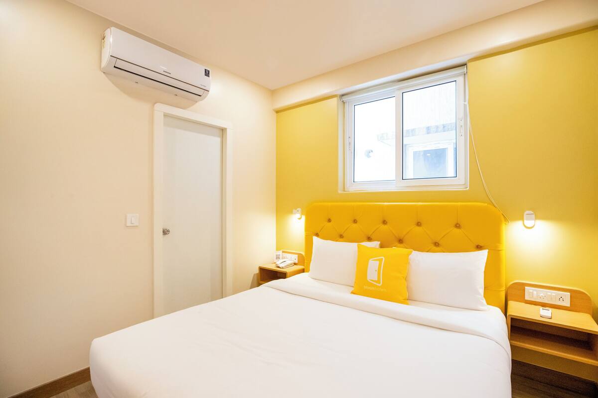 Quarto de hotel com cama, mesa de cabeceira, ar condicionado, janela e luminária na parede.