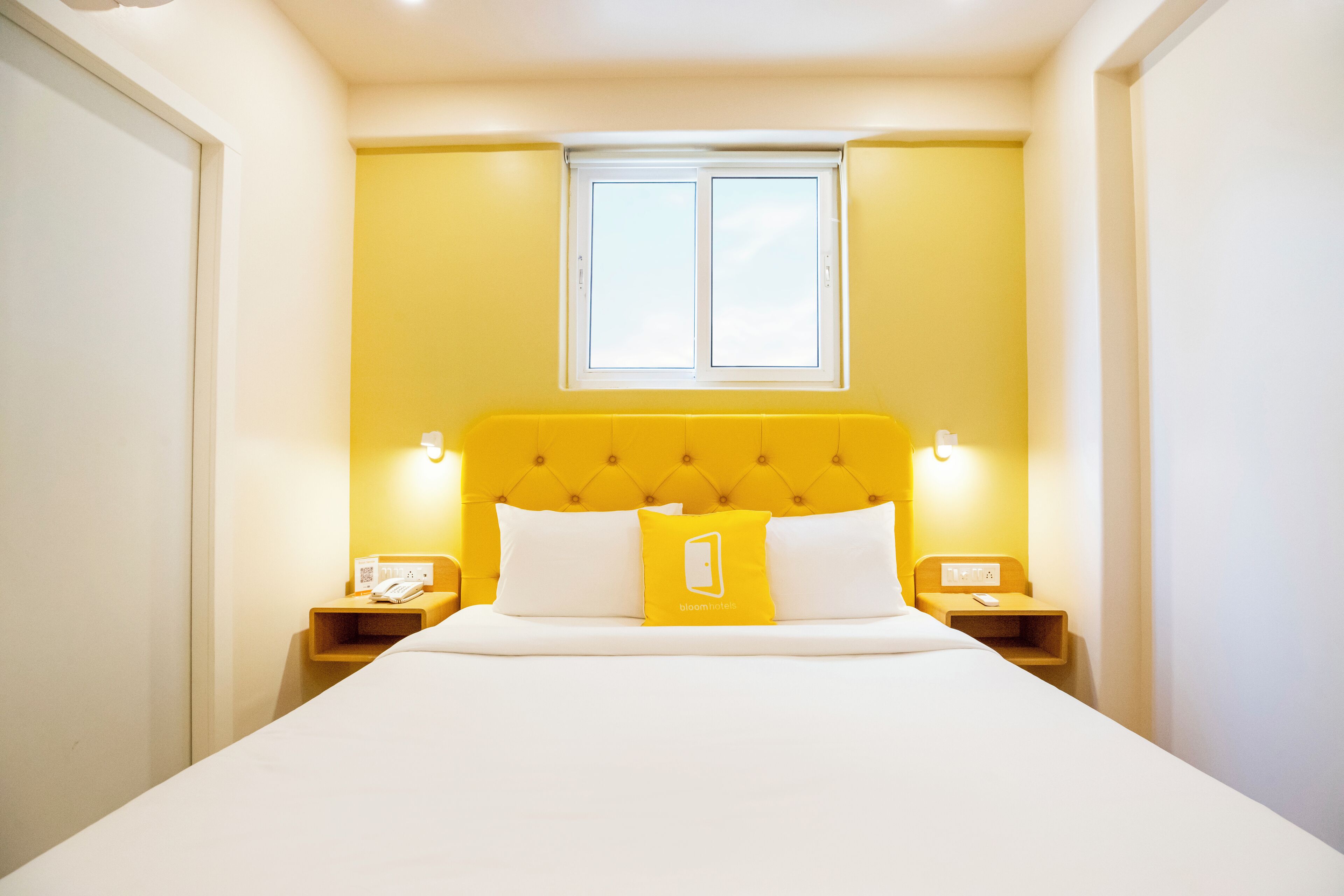 A hotel room with a yellow headboard, white bedding, and a small bedside table with a phone and a lamp.
