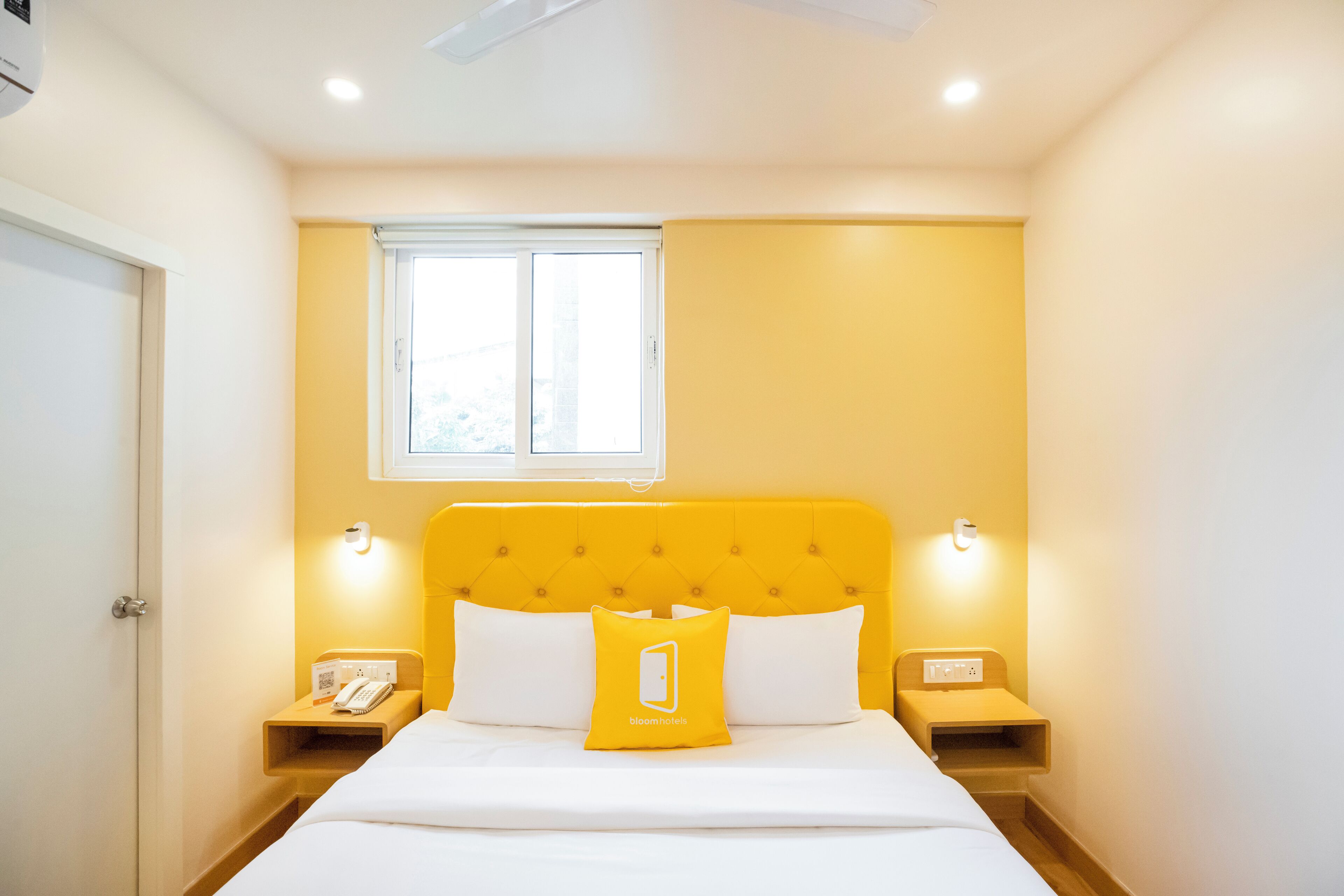 A hotel room with a yellow headboard, white bedding, a bedside table with a phone, and a window above the bed.