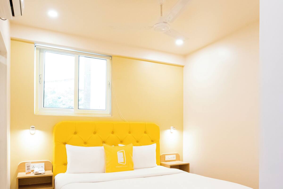Quarto de hotel com cabeceira amarela, roupa de cama branca, ventoinha no teto, janela e abajures laterais.
