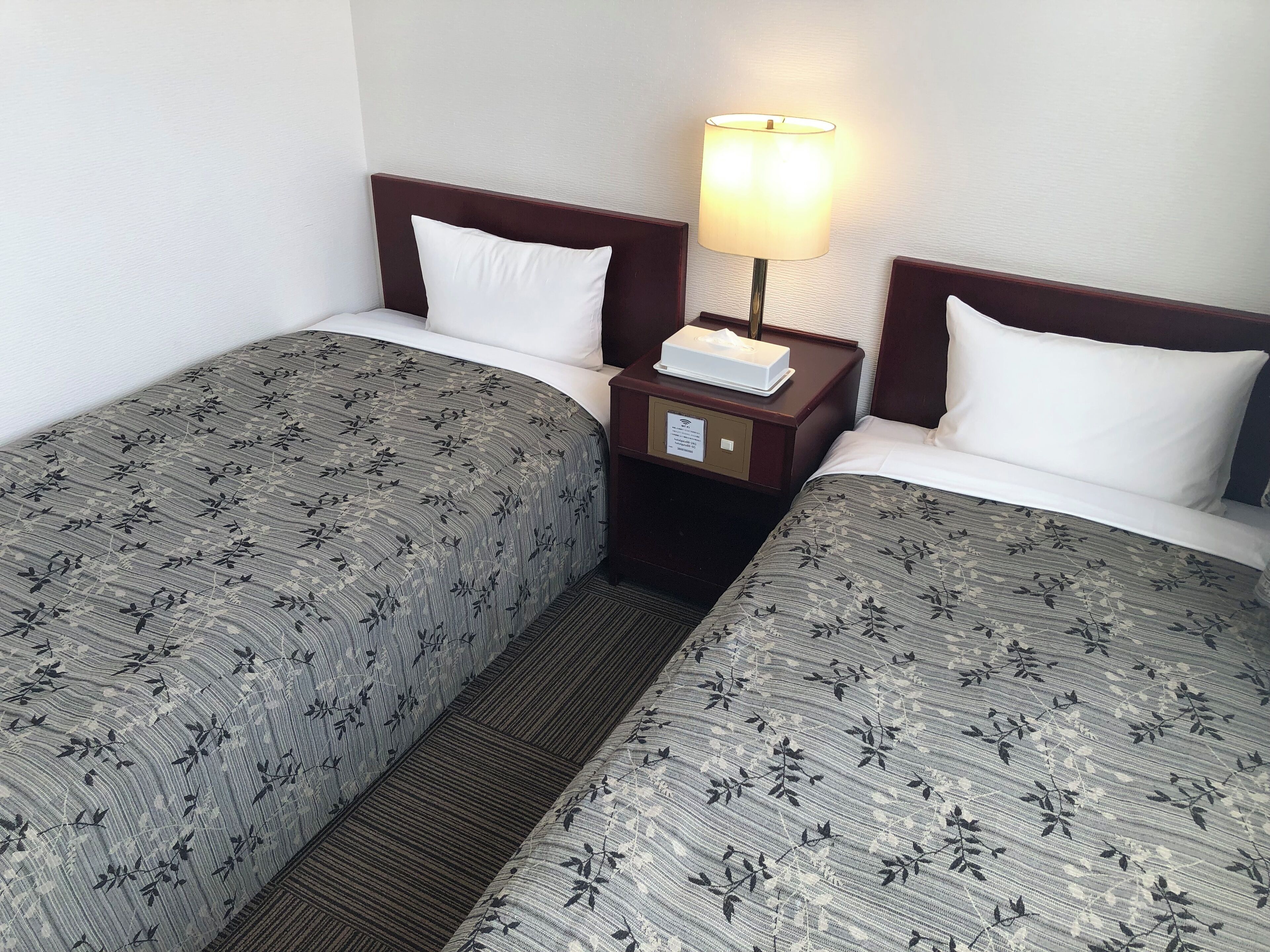 A hotel room with two single beds, each with a floral-patterned bedspread, a wooden nightstand with a lamp, and a small table with a tissue box.