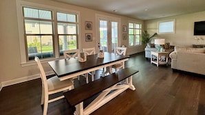 Un salon spacieux avec une grande table à manger en bois foncé, des chaises blanches et un canapé. De grandes fenêtres laissent entrer la lumière naturelle et offrent une vue sur l’extérieur.