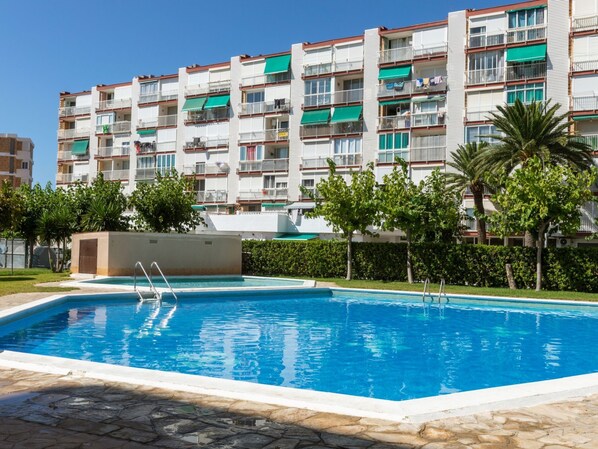 A swimming pool with a stone border, surrounded by a well-maintained lawn and trees. In the background, there is a multi-story residential building with balconies and green awnings. - Beach La Pineda by Interhome (La Pineda)