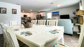 Un comedor con una mesa blanca dispuesta para seis personas, rodeada de sillas blancas. Una cesta de mimbre con objetos está sobre la mesa. En el fondo, se ve una cocina con armarios y electrodomésticos blancos.