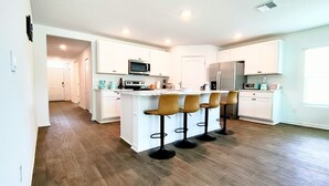 Una cocina moderna con armarios blancos, una isla central con taburetes y electrodomésticos como microondas y refrigerador.