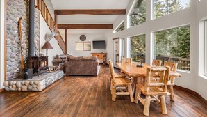 A spacious living area with a stone fireplace, wooden furniture, and large windows offering a view of the outdoors. - A-Frame for 16 with Hot Tub & Sauna (Cle Elum)