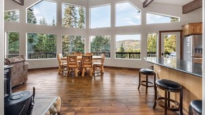 A spacious room with a high ceiling, large windows, a wooden dining table with chairs, a kitchen area, and a cozy seating area with a sofa and a fireplace. - A-Frame for 16 with Hot Tub & Sauna (Cle Elum)