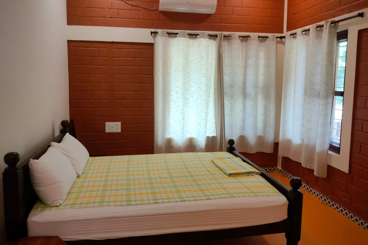 A bedroom with a checkered bedspread, a wooden headboard, white curtains, and a brick wall.