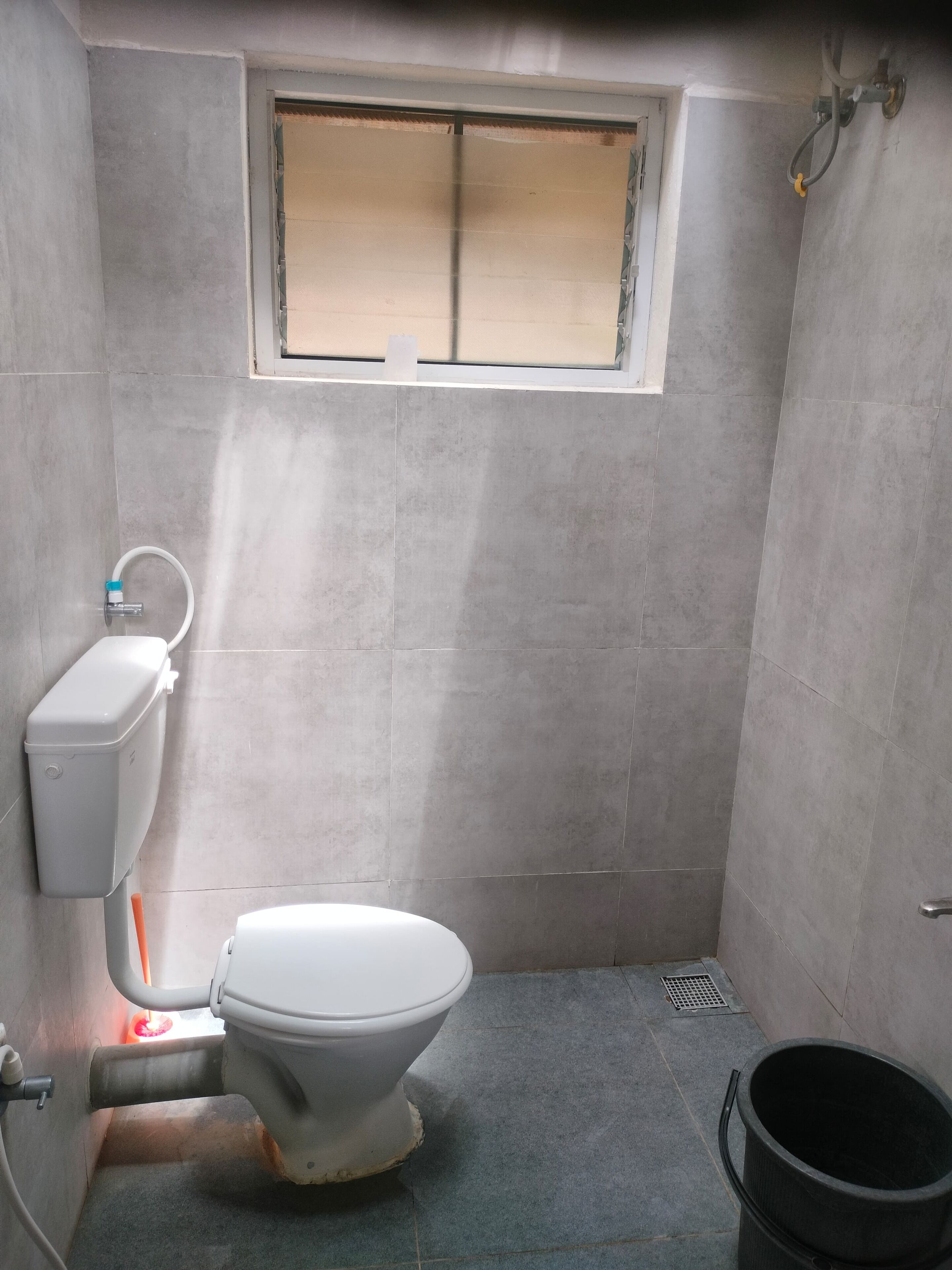 Ein kleines Badezimmer mit Fliesen, einer Toilette, einem Fenster und einem schwarzen Eimer.