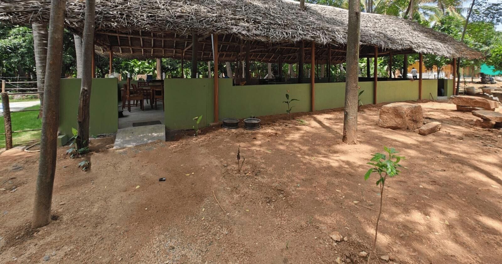 A rustic thatched-roof structure with a green exterior, surrounded by trees and a dirt ground.