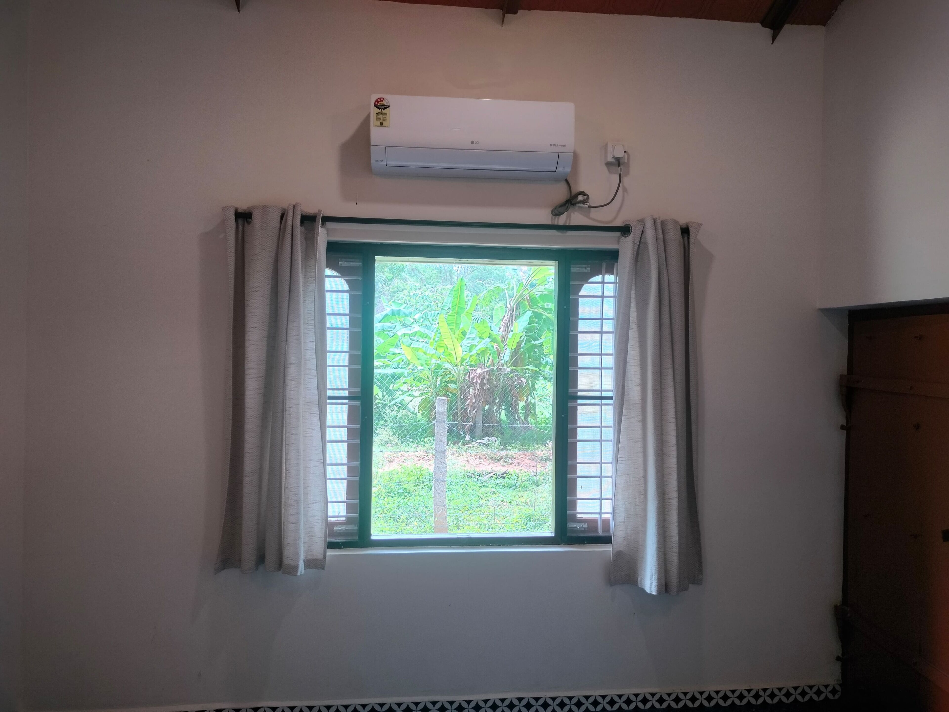 A room with a window, an air conditioning unit above it, and a view of greenery outside.