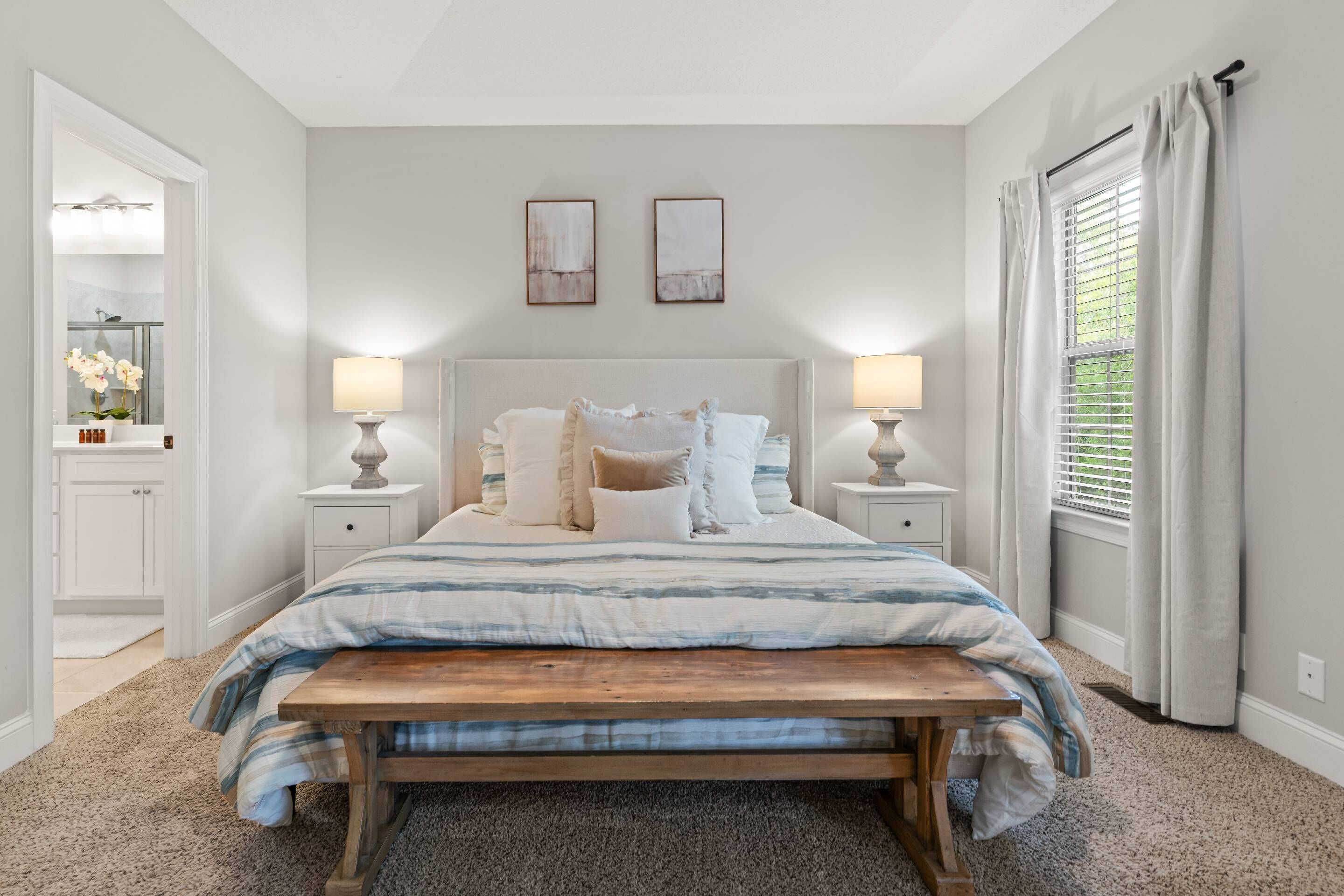 A bedroom with a large bed, two bedside lamps, a bench, a nightstand, a mirror, and a window with curtains.