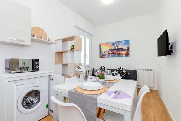 A compact living space with a washing machine, a dining table with chairs, a TV mounted on the wall, and a painting above the table.