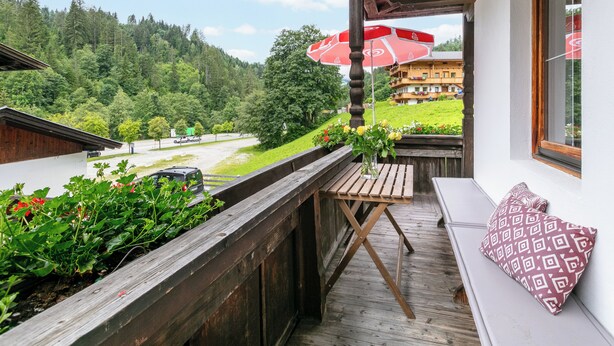 A balcony with a wooden table, a red umbrella, and a view of a mountain and a building in the background.