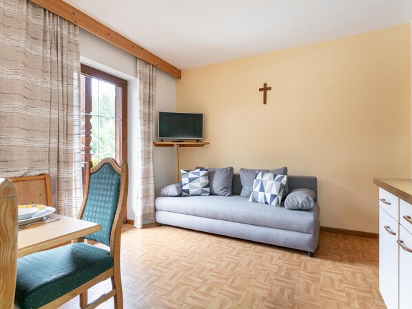 A cozy living room with a sofa, a television mounted on a wooden stand, a cross on the wall, and a dining area with a table and chairs.
