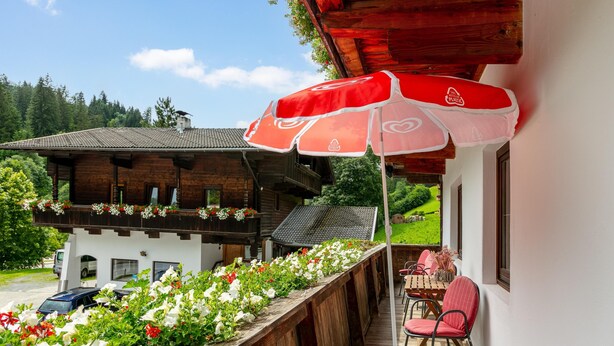 Un balcon en bois avec un parasol rouge, une table et des chaises, et un parterre de fleurs.