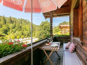 A wooden balcony with a table set for dining, overlooking a forested valley and a building with balconies.