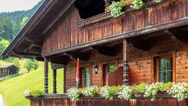 A wooden chalet with a balcony adorned with flowers, a wooden porch, and a mountain backdrop.