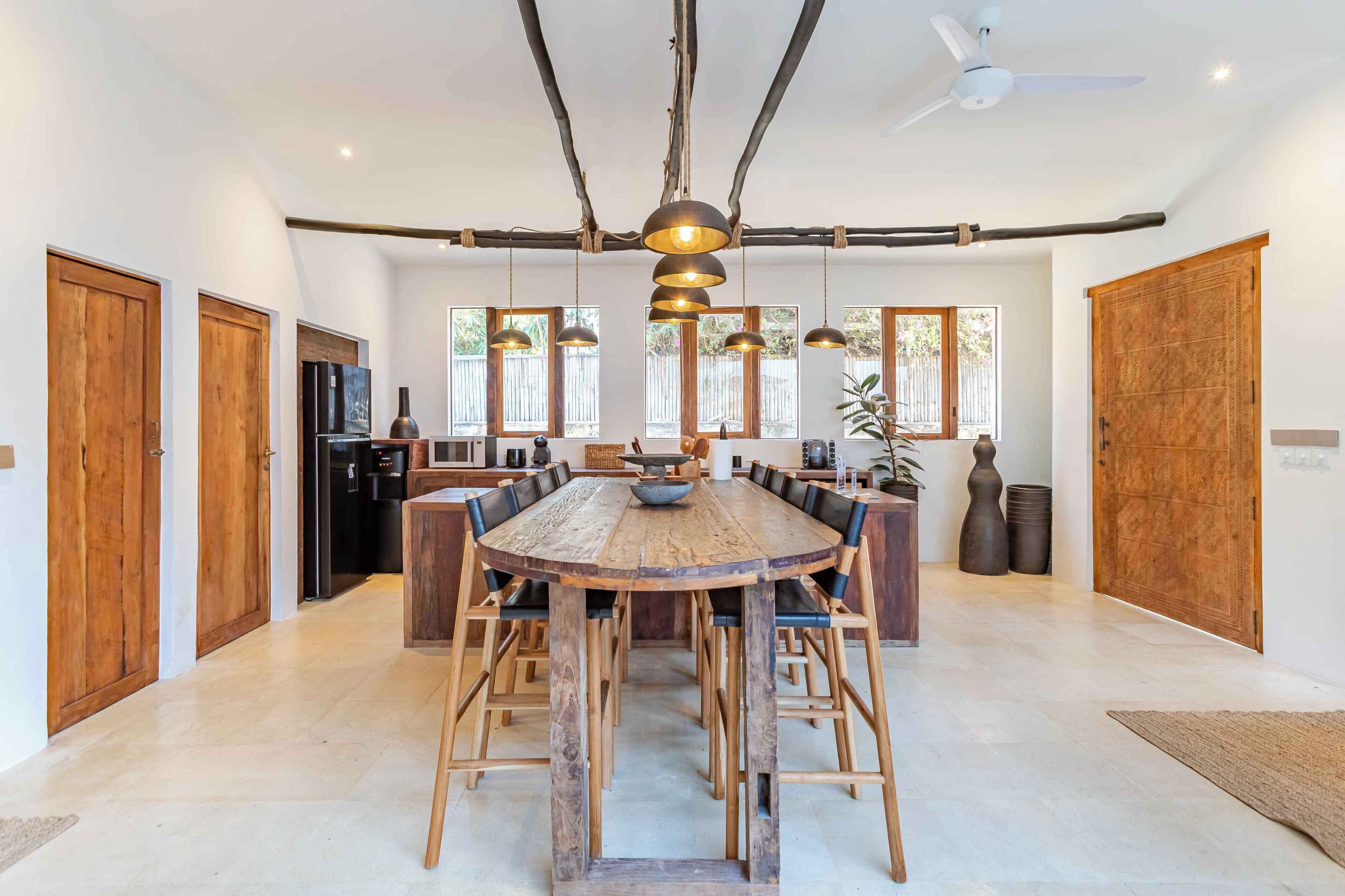 Uma ampla área de jantar com uma grande mesa de madeira e cadeiras, uma cozinha moderna ao fundo e grandes janelas que proporcionam luz natural.