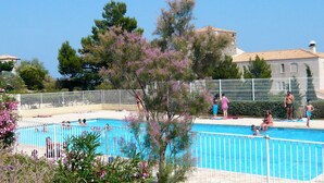 Una piscina rodeada por una cerca blanca, con gente nadando y permaneciendo cerca de la piscina. Se ven árboles y un edificio al fondo.