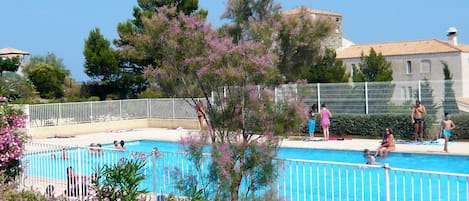 A swimming pool surrounded by a white fence, with people swimming and standing by the pool. There are trees and a building in the background.
