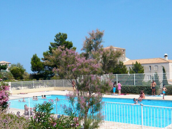 A swimming pool surrounded by a white fence, with people swimming and standing by the pool. There are trees and a building in the background.